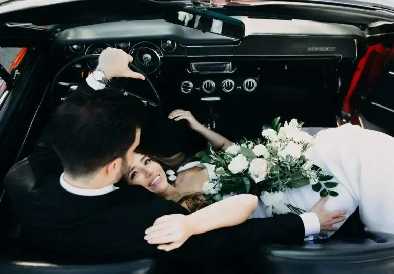 Posed for a photo, a groom pretends to drive a convertible while the bride lays on the seat next to him with her head in his lap, smiling at the camera above.