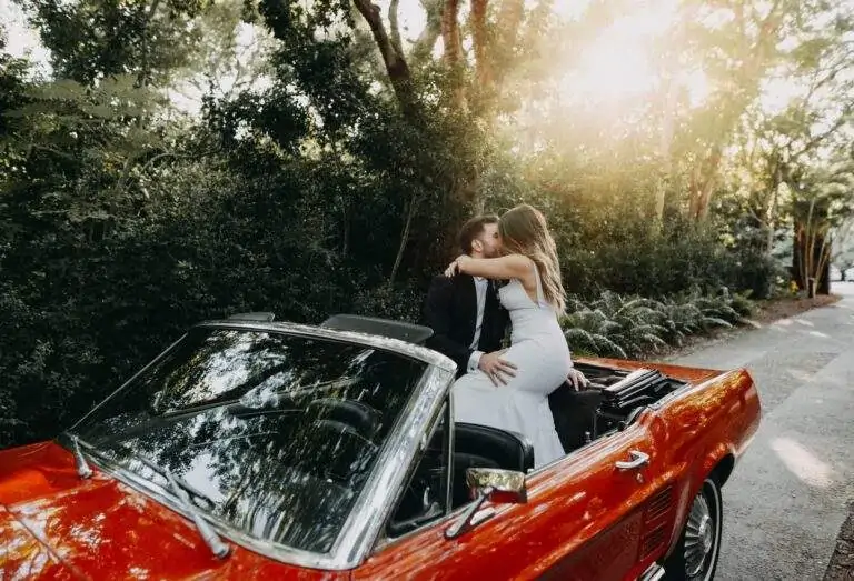 A bride and groom sit atop a red convertible as they kiss.