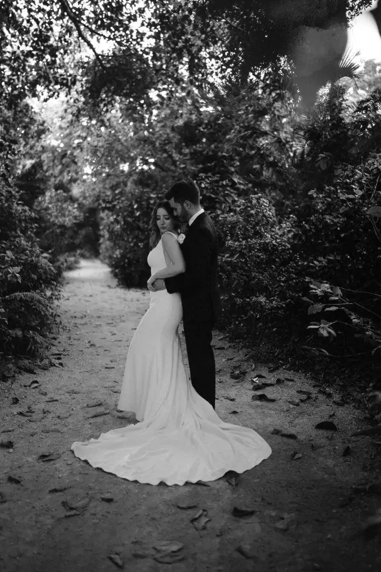 In black and white, a groom stands behind a bride with his arms around her in embrace as they pose for a photo.
