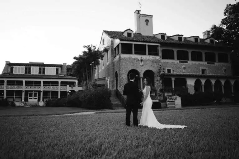 In black and white, a bride and groom stand on the main lawn of Deering Estate facing away from the camera toward the Richmond Cottage and Stone House.