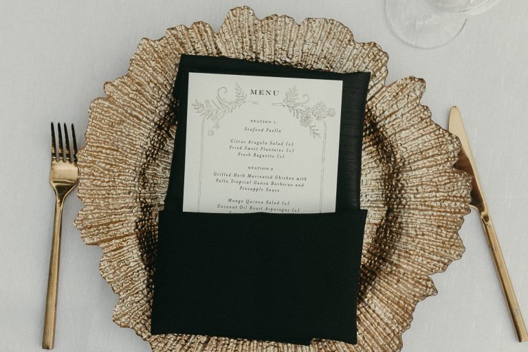 Detail of the table featuring a gold coastal-inspired charger plate, gold fork and knife, and a black folded napkin with a wedding menu tucked neatly inside