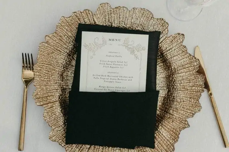 Detail of the table featuring a gold coastal-inspired charger plate, gold fork and knife, and a black folded napkin with a wedding menu tucked neatly inside