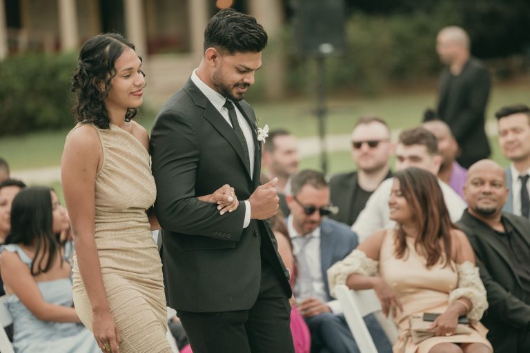 Two members of the wedding party, one man and one woman, walking down the aisle towards the ceremony arch with seated guests in the background