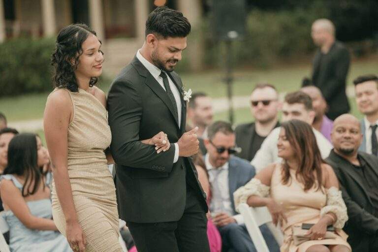 Two members of the wedding party, one man and one woman, walking down the aisle towards the ceremony arch with seated guests in the background