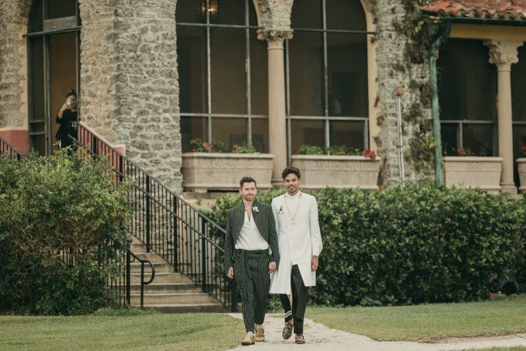 Both grooms walking hand-in-hand towards their wedding arch for their ceremony. The historical Stone House Museum is in the background.