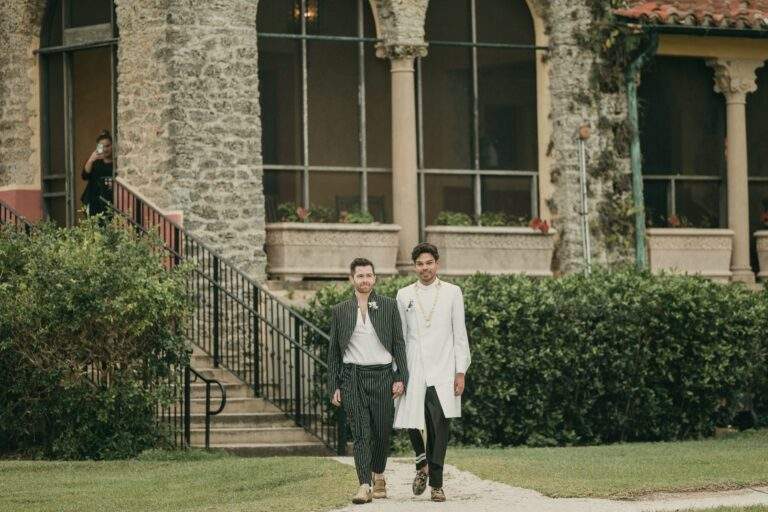 Both grooms walking hand-in-hand towards their wedding arch for their ceremony. The historical Stone House Museum is in the background.