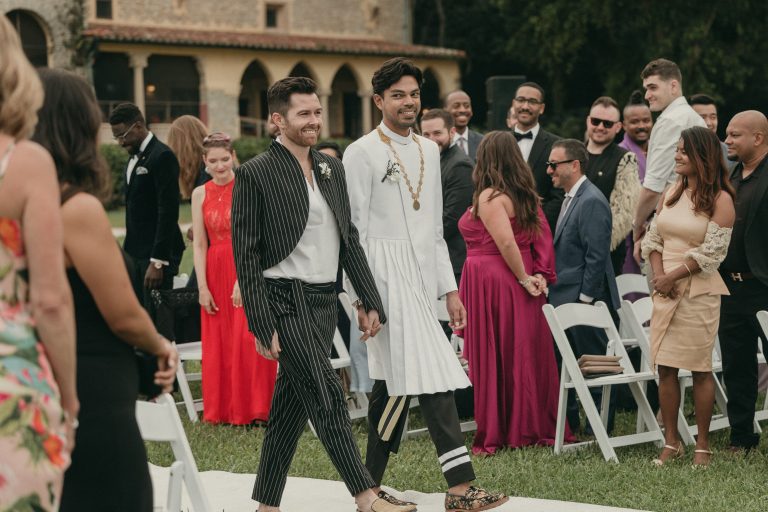 Both grooms walking hand-in-hand towards their wedding arch for their ceremony. Guests are standing and smiling in the background.