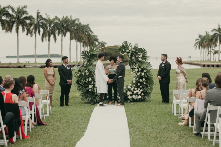 Grooms holding hands during their waterfront wedding ceremony at Deering Estate