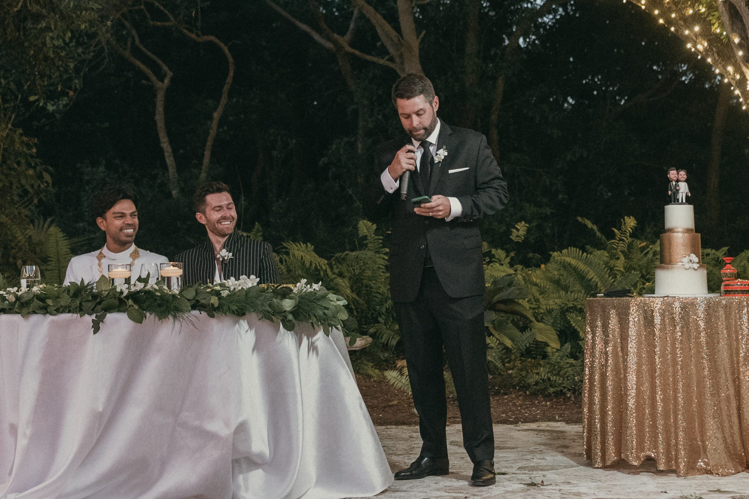 A guest giving a wedding speech at the historic courtyard while the grooms sit at the sweetheart's table to the guests' left. A wedding cake on a cake table is visible to the right,