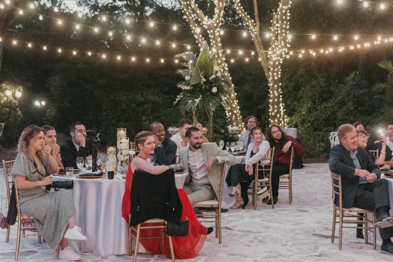 Wedding guests seated at their tables intently listening to wedding speeches. Bistro lights glow above them.