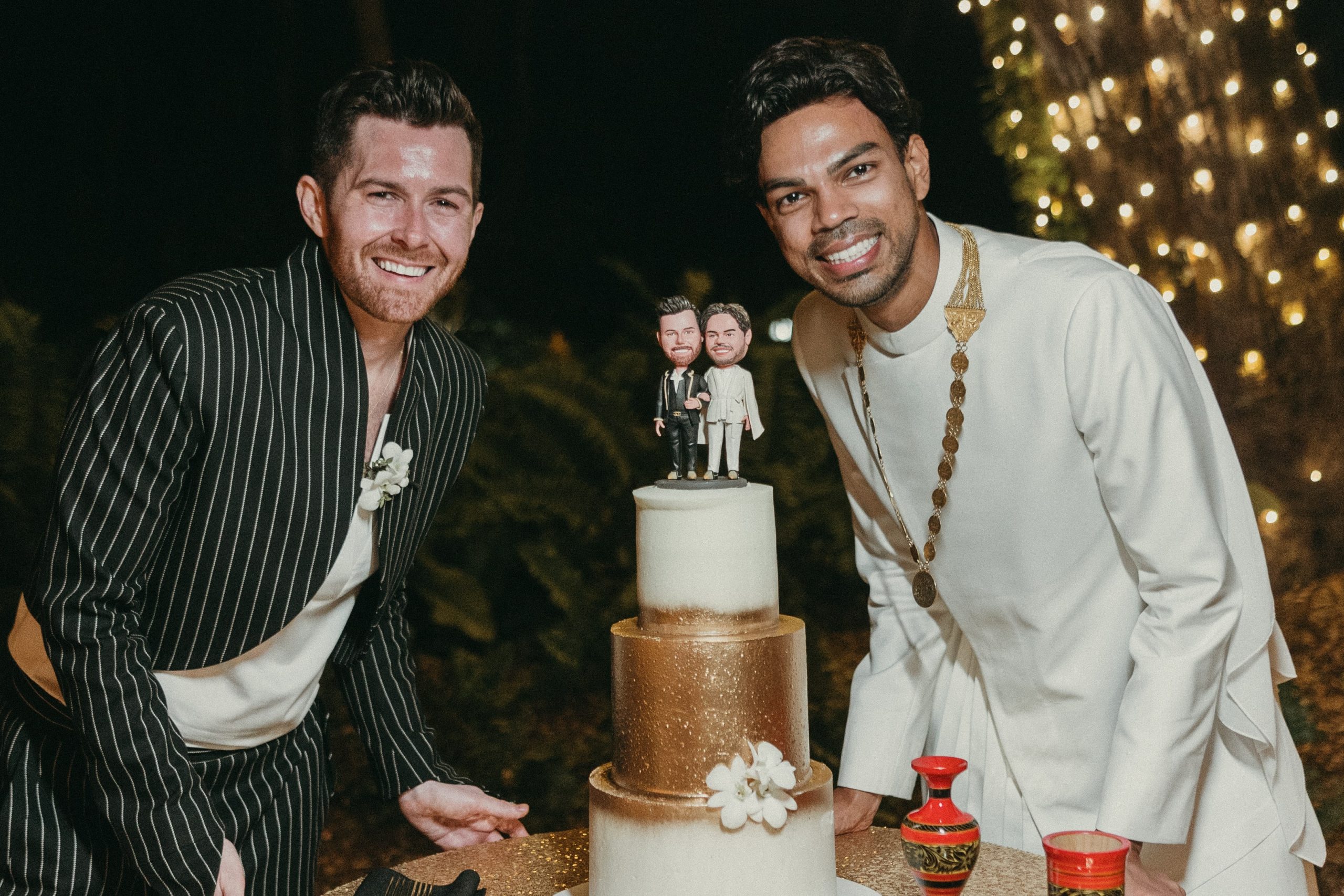Grooms happily posing next to their wedding cake, topped with two bobble heads that are modeled after them