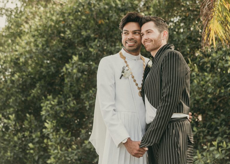 Grooms posing for a picture while embracing each other and holding hands with Deering Estate's tropical gardens in the backdrop