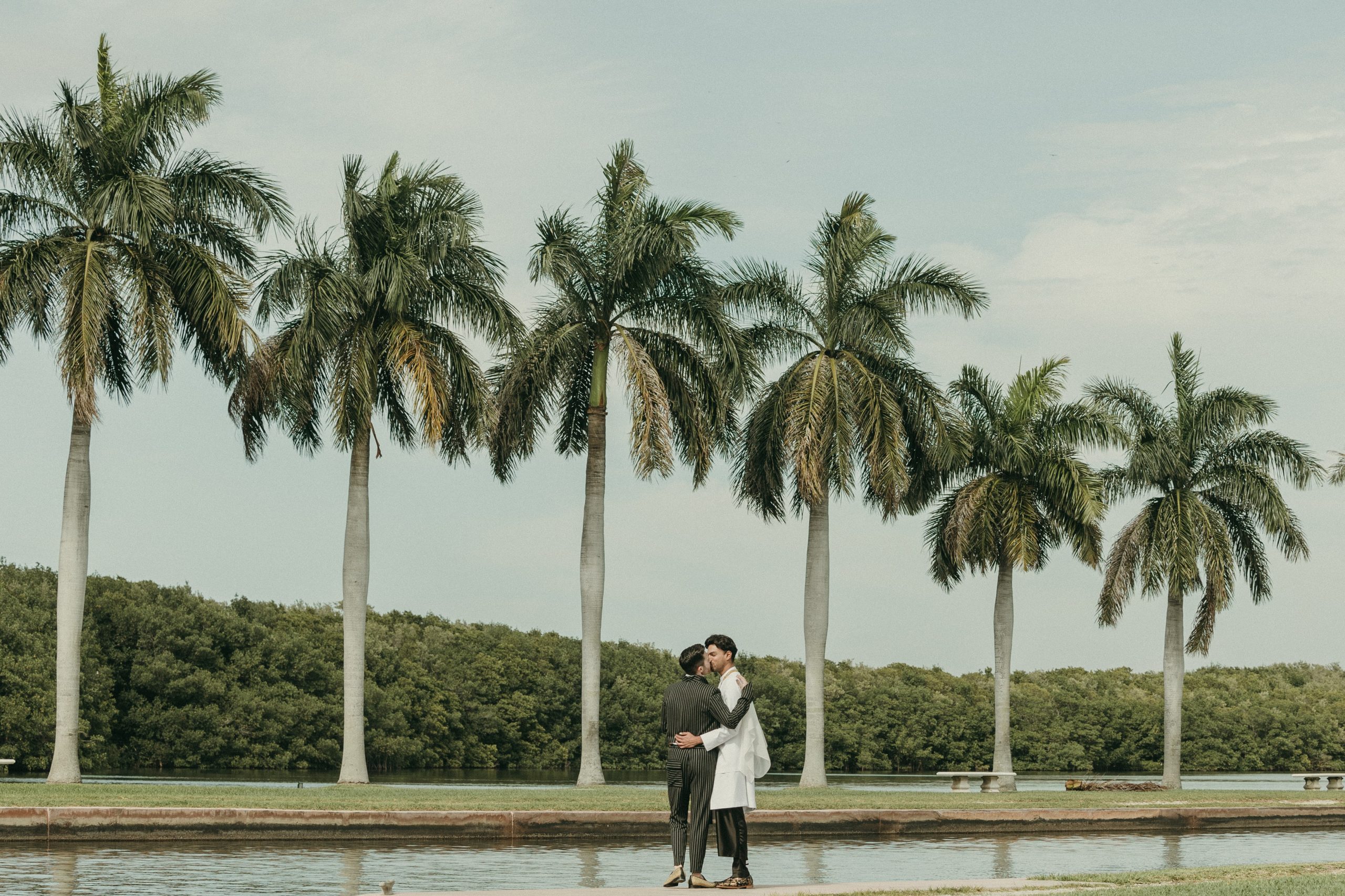 Grooms embracing each other while at Deering Estate's waterfront lawn. Biscayne Bay and six palm trees are in the background, and mangrove trees are visible behind the palm trees.