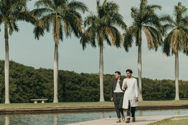 Grooms walking towards the camera at Deering Estate's waterfront hand-in-hand with palm trees and Biscayne Bay in the background