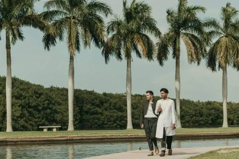 Grooms walking towards the camera at Deering Estate's waterfront hand-in-hand with palm trees and Biscayne Bay in the background