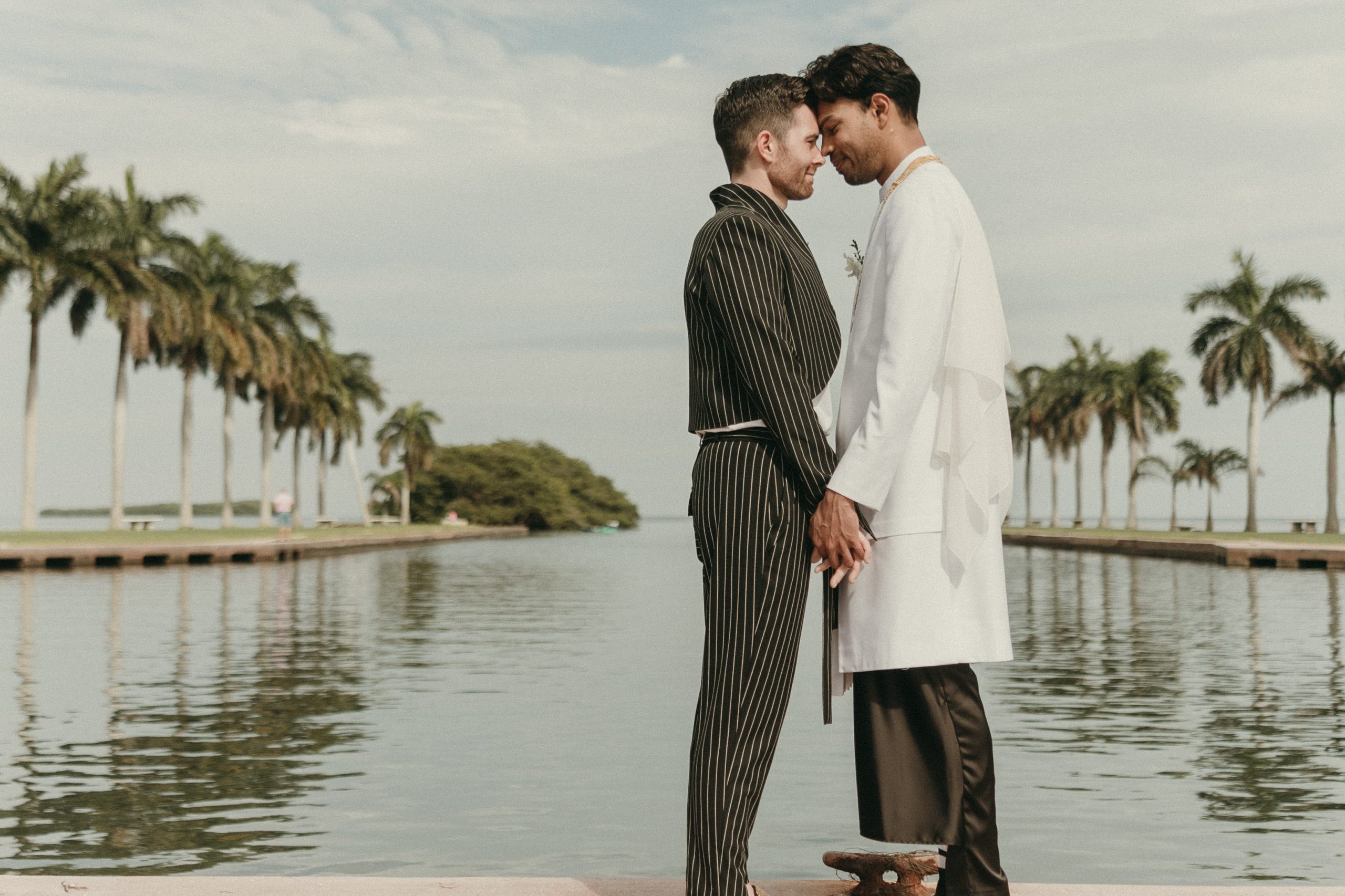Grooms embracing each other at Deering Estate's waterfront boat basin. Biscayne Bay is in the background, and rows of palm trees line both sides of the photo.
