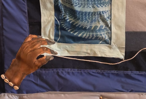 A close-up view of a textile artwork featuring a painted hand holding a thin cord. The hand is crafted from fabric and attached to the larger textile piece, with several small shell-like beads sewn near the wrist. Surrounding the hand are stitched fabric panels in shades of blue, cream, and gray. One panel contains a printed or woven circular blue pattern resembling basketry.