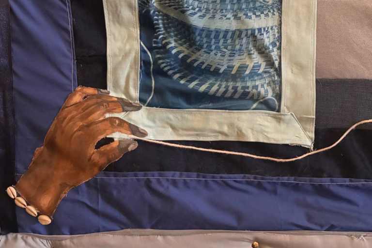 A close-up view of a textile artwork featuring a painted hand holding a thin cord. The hand is crafted from fabric and attached to the larger textile piece, with several small shell-like beads sewn near the wrist. Surrounding the hand are stitched fabric panels in shades of blue, cream, and gray. One panel contains a printed or woven circular blue pattern resembling basketry.