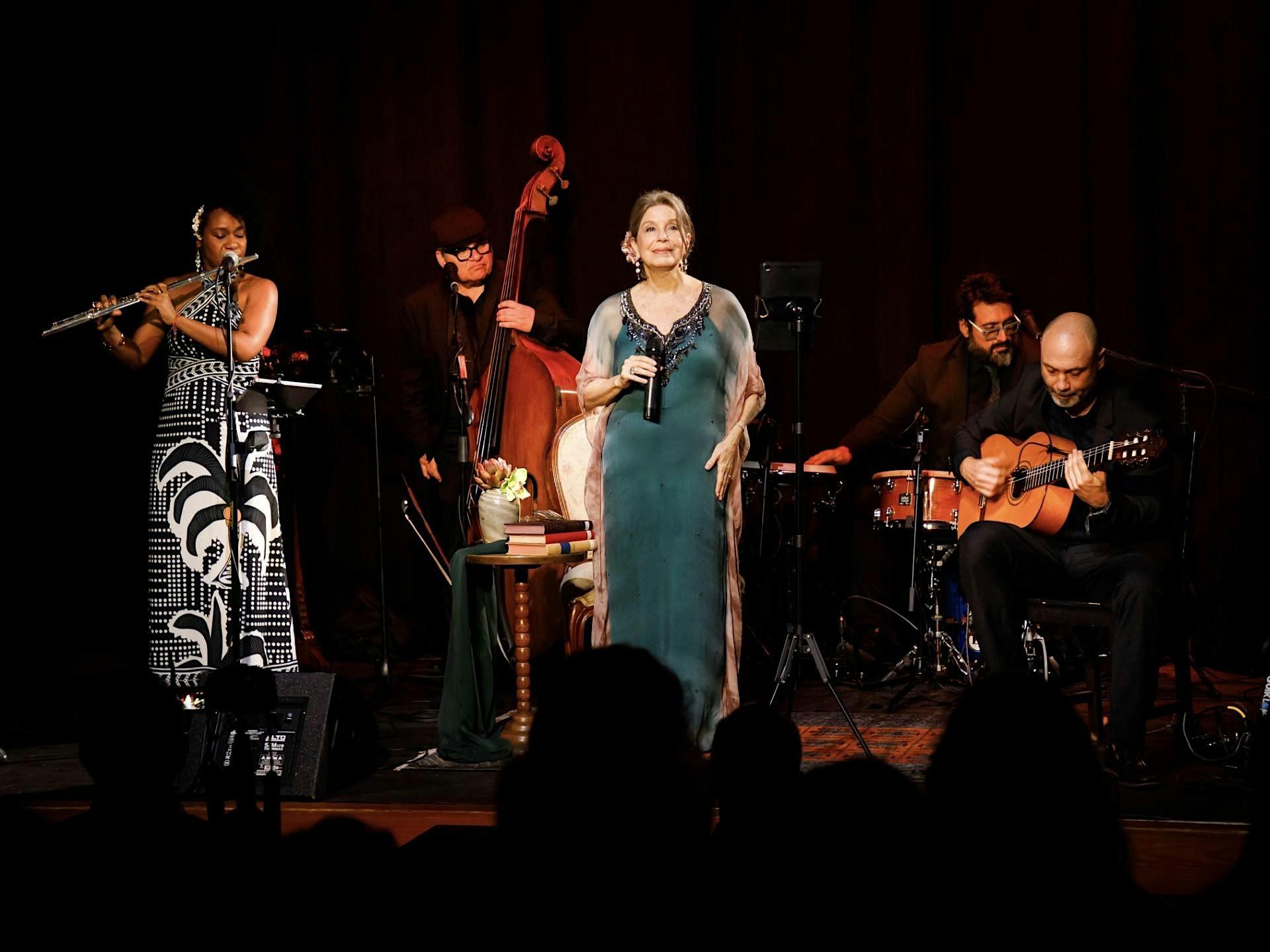 A woman in a black and white dress playing the flute, a man playing the standing bass, a man playing the drums, a man playing an acoustic guitar, and a woman in a green dress singing into a microphone on stage.