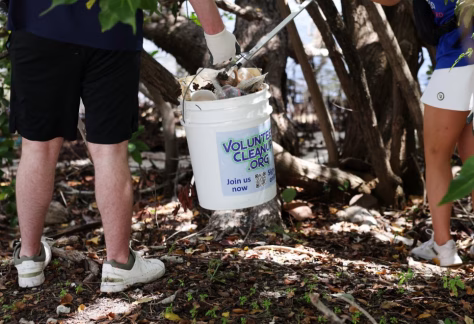Two people collect trash in a wooded area near the water, placing debris into a white bucket labeled “VolunteerCleanup.org.”