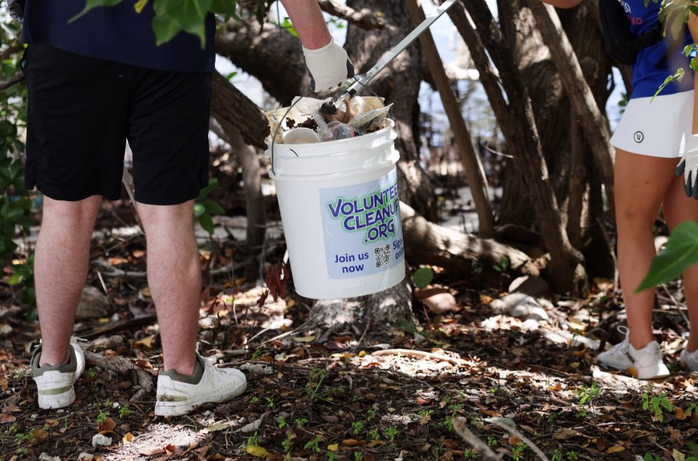 Two people collect trash in a wooded area near the water, placing debris into a white bucket labeled “VolunteerCleanup.org.”