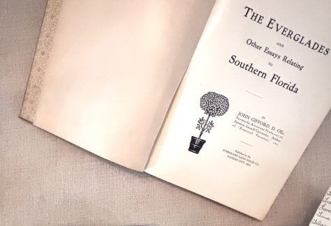 An open book rests on a light, textured fabric surface. The left page is blank, while the right page shows a title page with printed text reading “The Everglades and Other Essays Relating to Southern Florida.” Below the title is a small black illustration of a potted, round-canopied tree. Additional text on the page provides the author’s name and publishing information in smaller print. In the lower-right corner of the image, a separate small piece of paper with handwritten notes is partially visible.