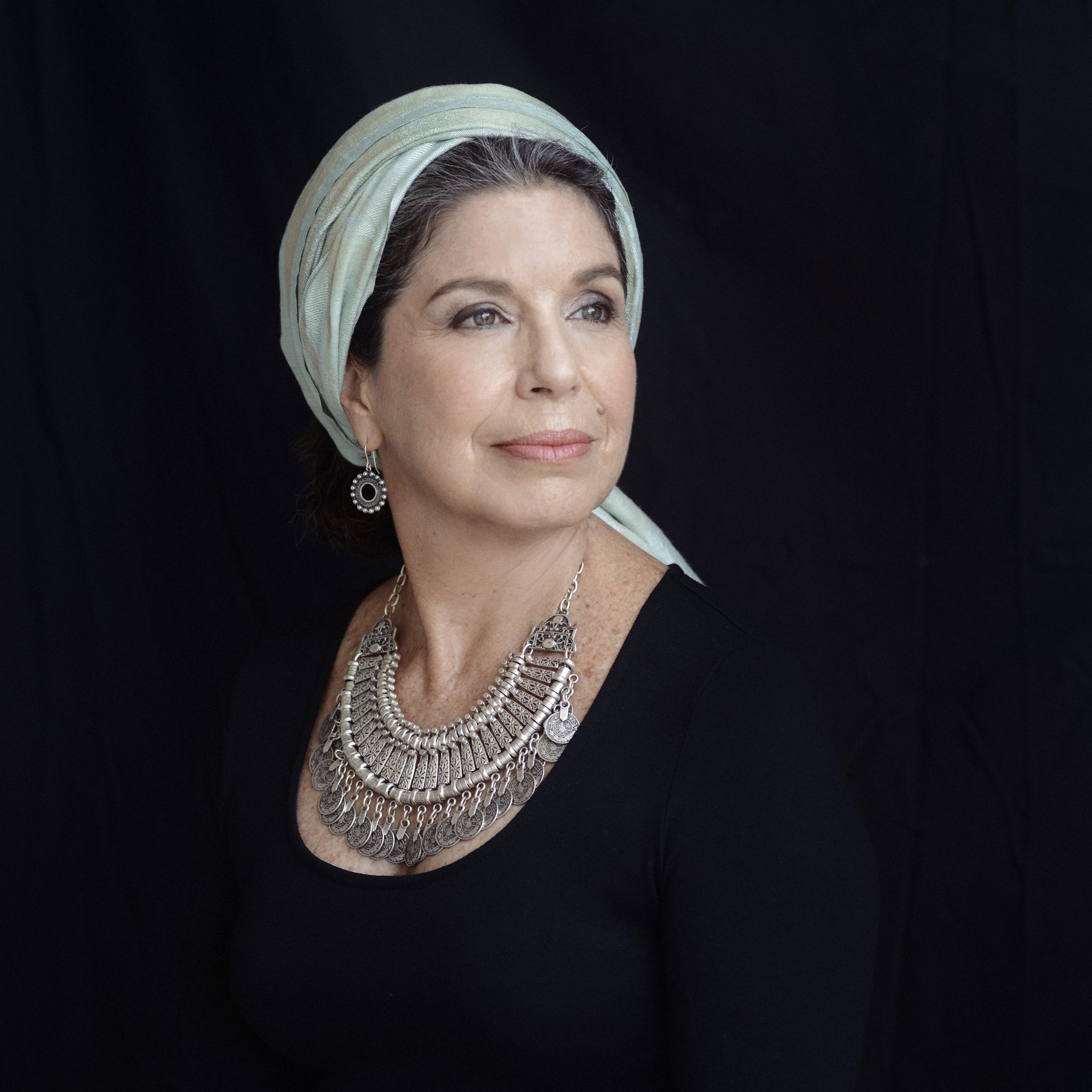 A person is posed against a dark backdrop, wearing a light-colored headscarf wrapped around the head, a dark long‑sleeved top, a large layered metallic necklace with ornate detailing, and a dangling circular earring. The lighting highlights the accessories and upper torso.