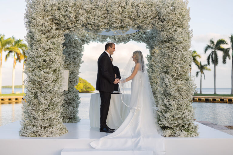 A groom and bride hold hands and look lovingly at one another at the altar of their wedding ceremony in front of a large floral arch.