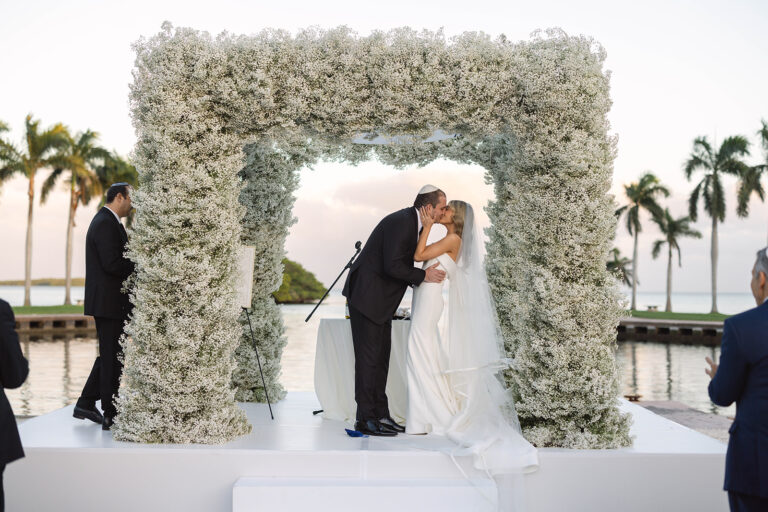 A bride and groom kissing at the altar of their wedding ceremony in front of a large floral arch.