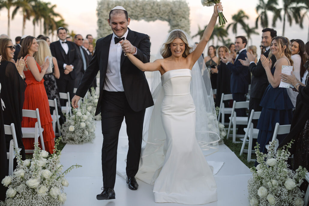 A bride and groom joyfully strut down the aisle together during their wedding ceremony, surrounded by floral decorations and their loved ones.
