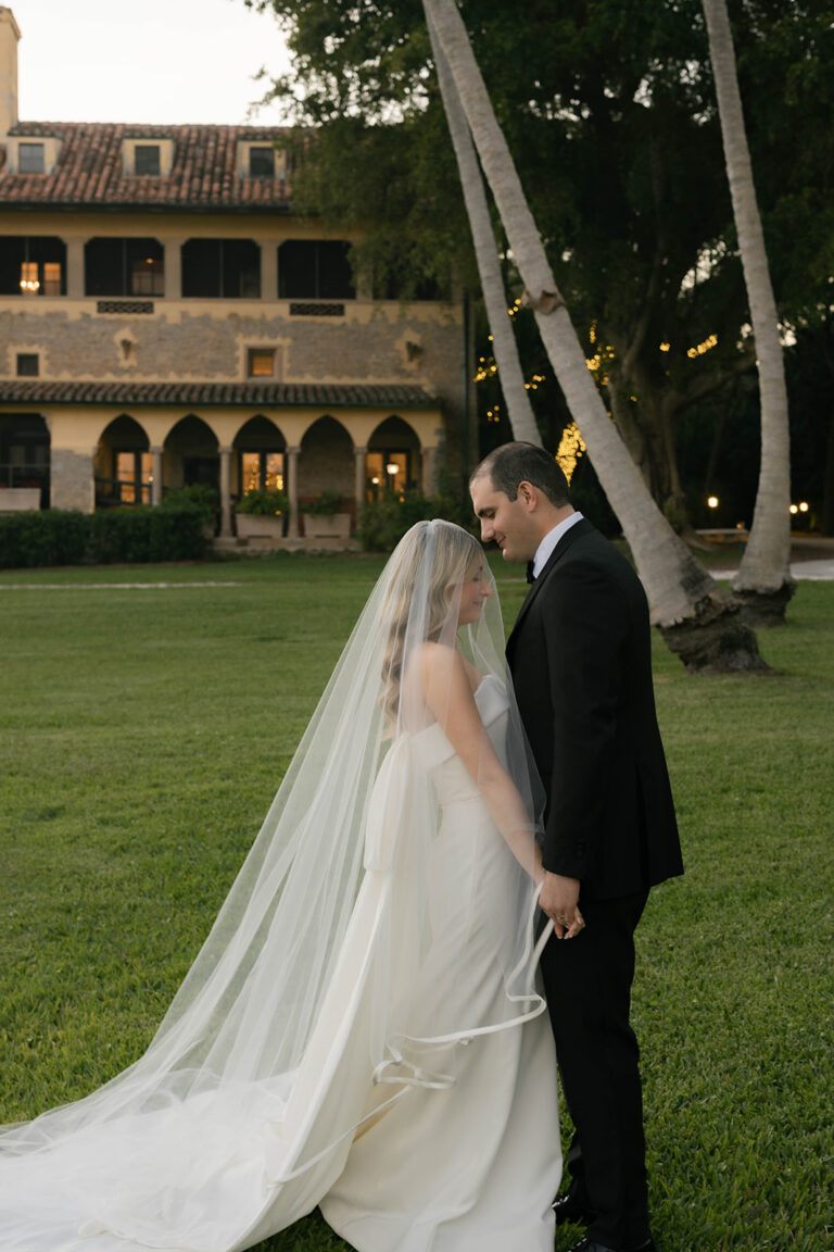 A bride in a long white wedding dress and long white veil stands holding hands with her groom wearing a black tux. They are facing each other with their eyes closed in a serence moment in front of the Stone House at Deering Estate.