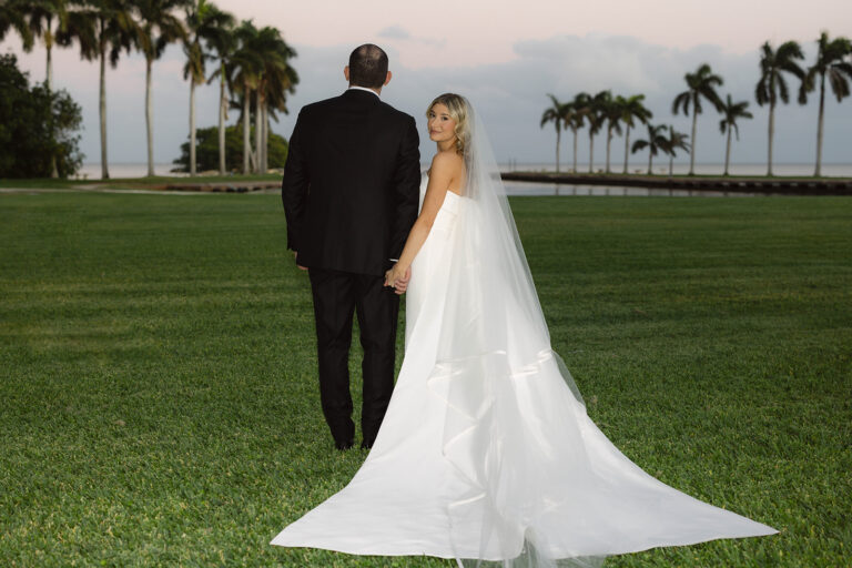A photo of a groom and bride on the Main Lawn at Deering Estate with a view of Biscayne Bay. The groom, wearing a black tux, is faced away from the camera looking out to the water, and the bride is looking back at the camera as she holds the groom's hand.