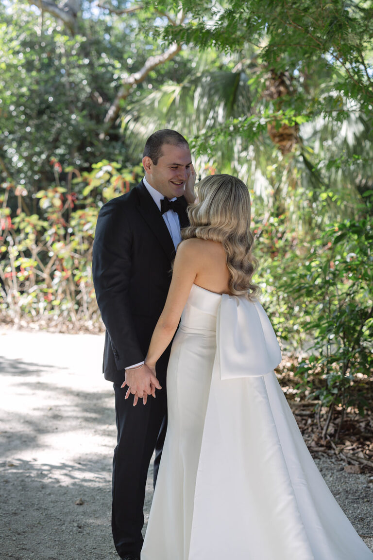 A groom smiles down at his bride. He is wearing a black tux. The bride is wearing a long white dress and is facing the groom. They are holding hands and the bride is cupping the groom's face.