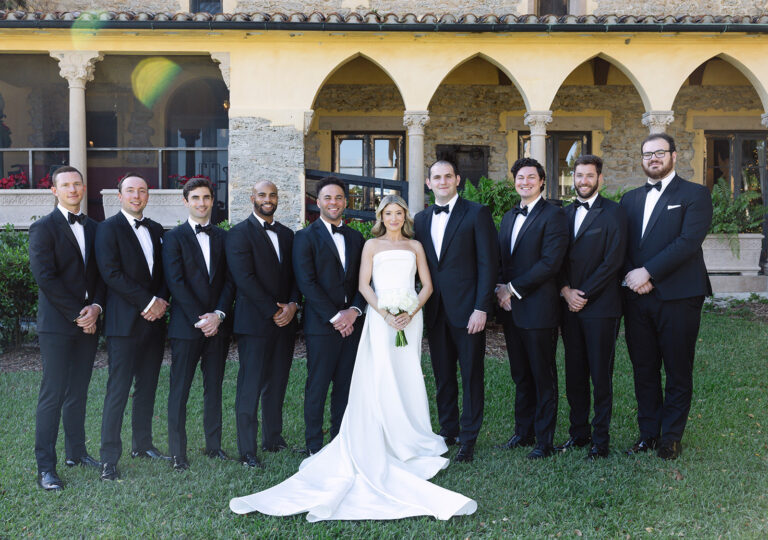 A bride in a long white dress holding a white boquet stands in the middle of nine groomsmen wearing black suits with bowties.