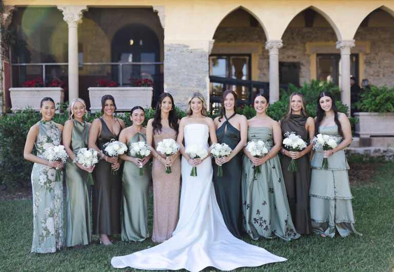 A bride in a long white dress stands in the middle of nine bridesmaids in green dresses with everyone holding a boquet of white flowers.