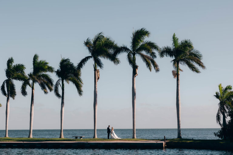 View of a bride and groom near the water on the jetty with palm trees at Deering Estate.