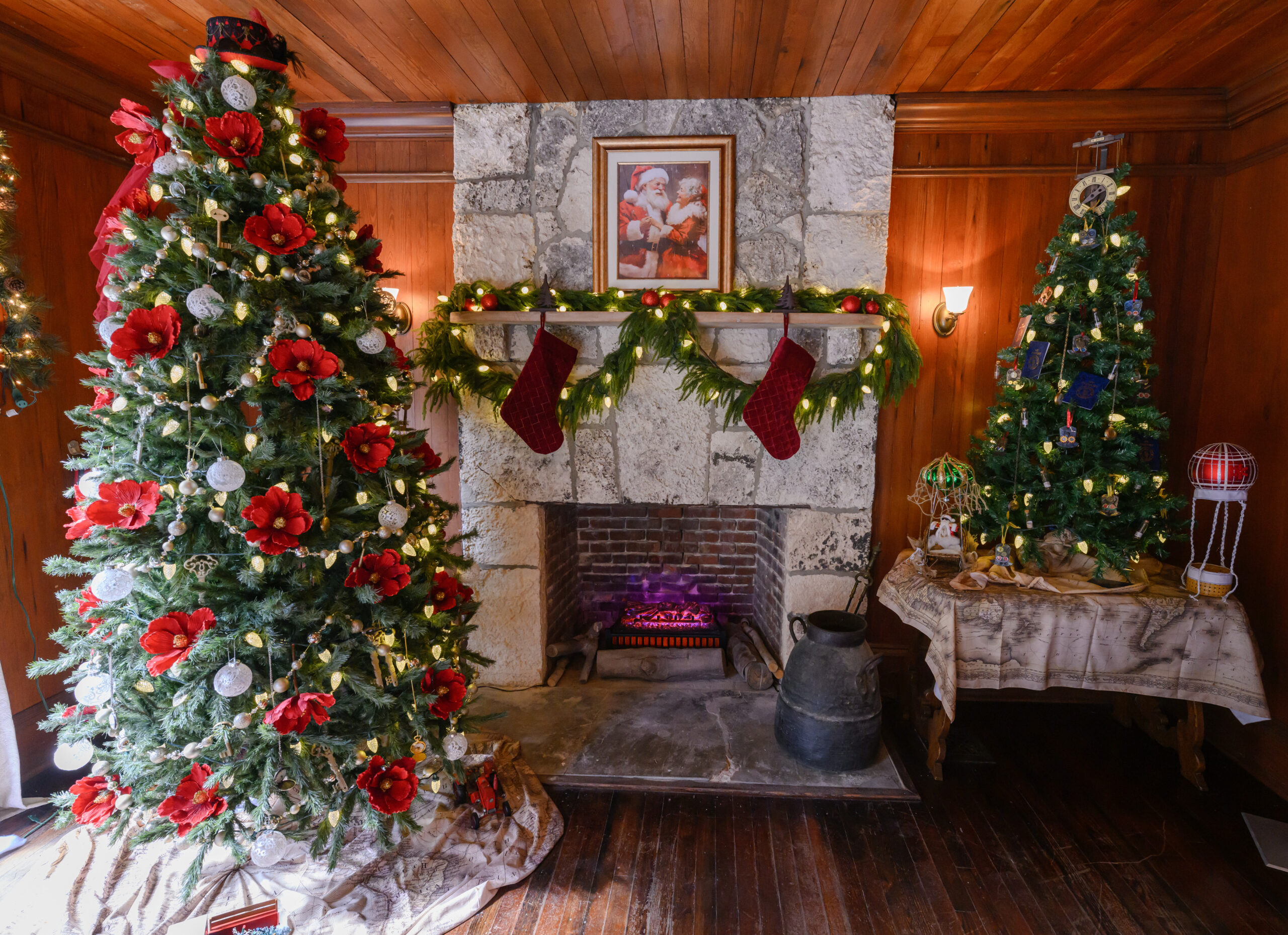 The fireplace area of the dining room in the Richmond Cottage decorated for the holidays. A photo of Mr. and Mrs. Clause hangs above the fireplace. To the left of the fireplace is a Christmas tree with poinsiettas and baubles. To the right of the fireplace is a smaller Christmas tree on a table with large white lights.