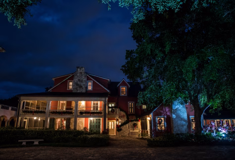 A historic two‑story house is illuminated at night with warm interior lighting and subtle holiday decorations. The building features red siding, stone chimneys, large porches, and green doors, surrounded by lush landscaping.