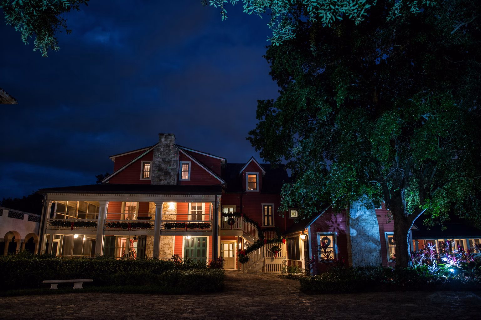 A historic two‑story house is illuminated at night with warm interior lighting and subtle holiday decorations. The building features red siding, stone chimneys, large porches, and green doors, surrounded by lush landscaping.