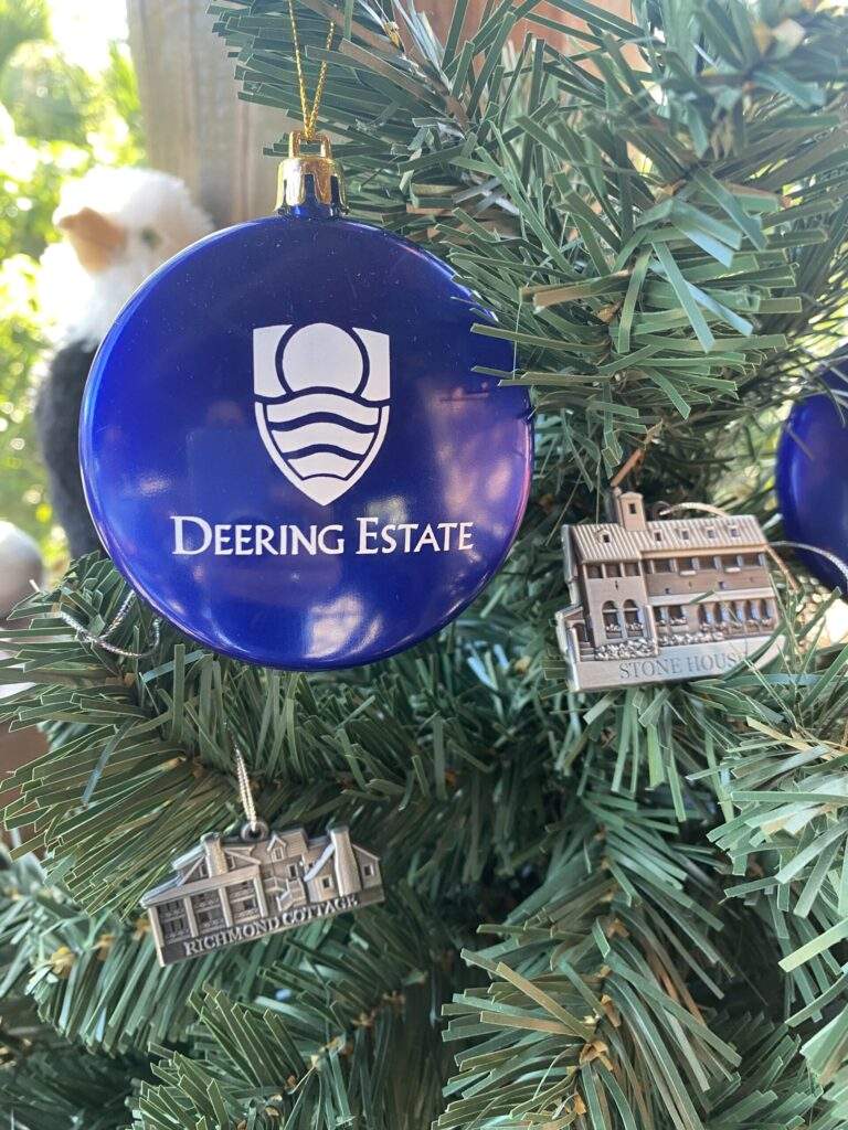 A blue circular ornament with the Deering Estate logo, an etched metal Richmond Cottage ornament, and an etched metal Stone House ornament displayed on a Christmas tree.