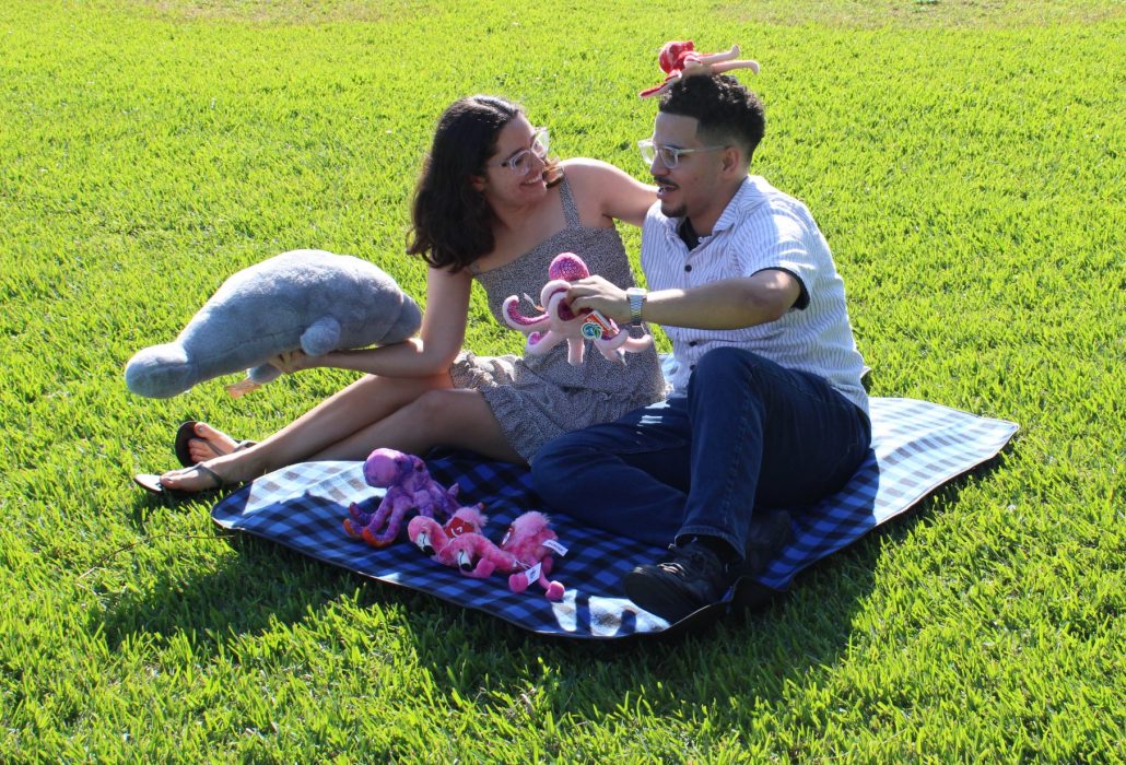 A couple sitting on a Deering Estate-branded picnic blanket on the Main Lawn playing with a manatee and octopus plushies.