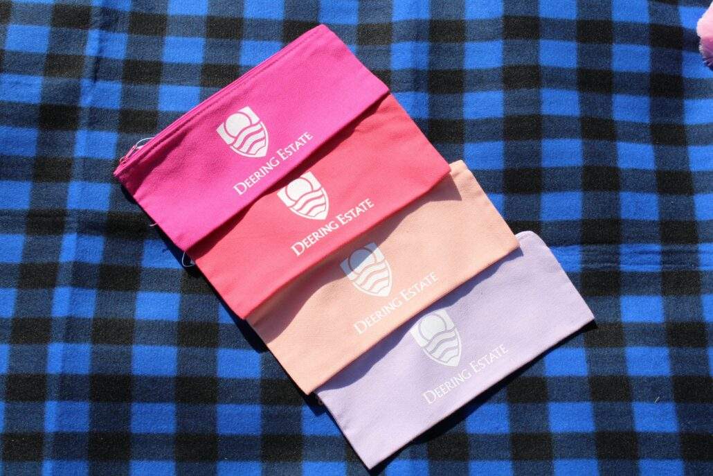 A pink, coral, peach, and light purple pencil pouch with the Deering Estate logo on them sit on a blue and black plaid picnic blanket.