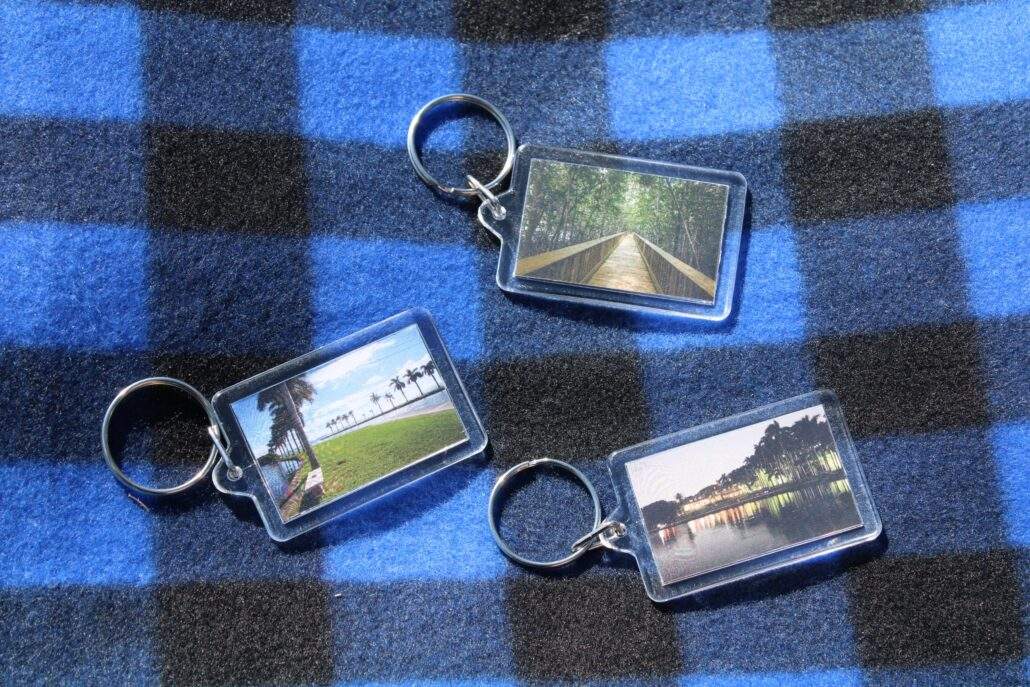 Three keychains featuring images of the Mangrove Boardwalk, North Jetty, and Boat Basin at Night sit on a blue and black plaid picnic blanket.