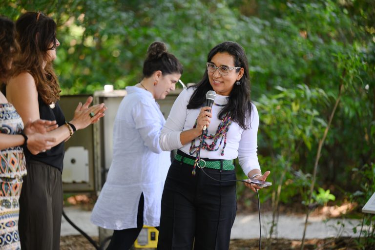 Curator Dainy Tapia. A woman with black hair, mulicolored glasses, white long-sleeve shirt, black pants, green belt, smiling and holding a microphone.