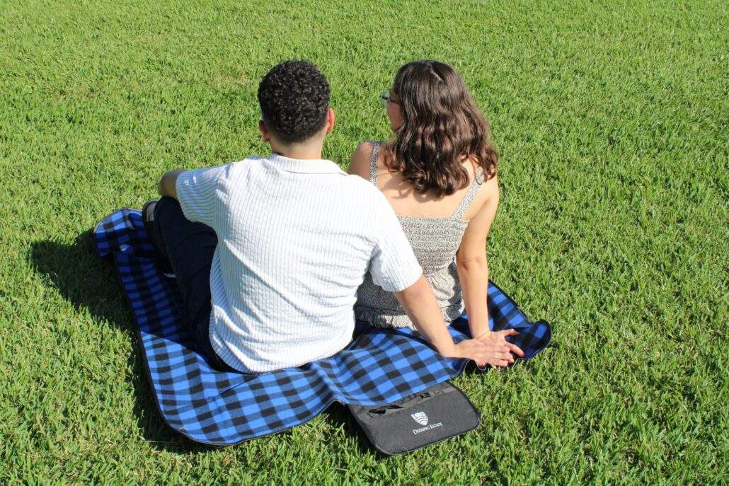 A young couple sitting together and holding hands on a Deering Estate-branded picnic blanket on the Main Lawn at Deering Estate.