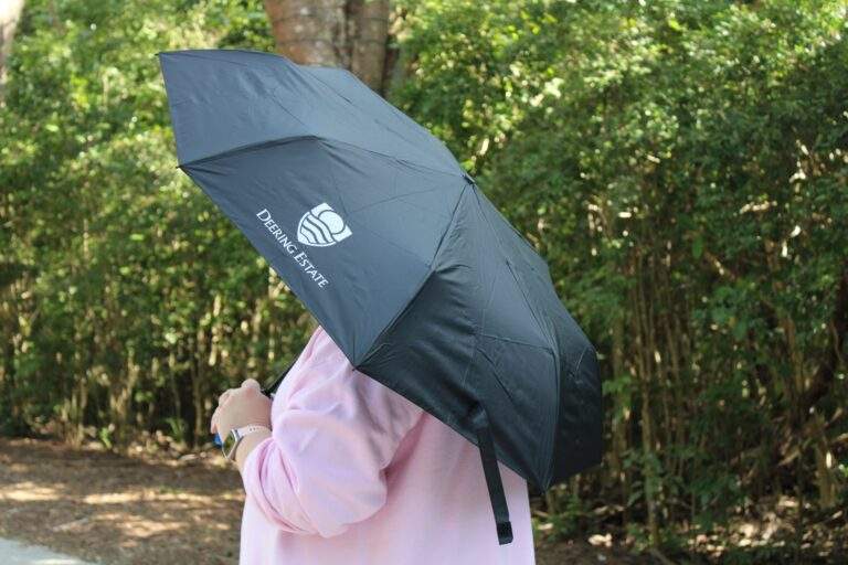 A person in a pink hoodie with their face obscured holding a black umbrella with the Deering Estate logo on it in front of green hedges.