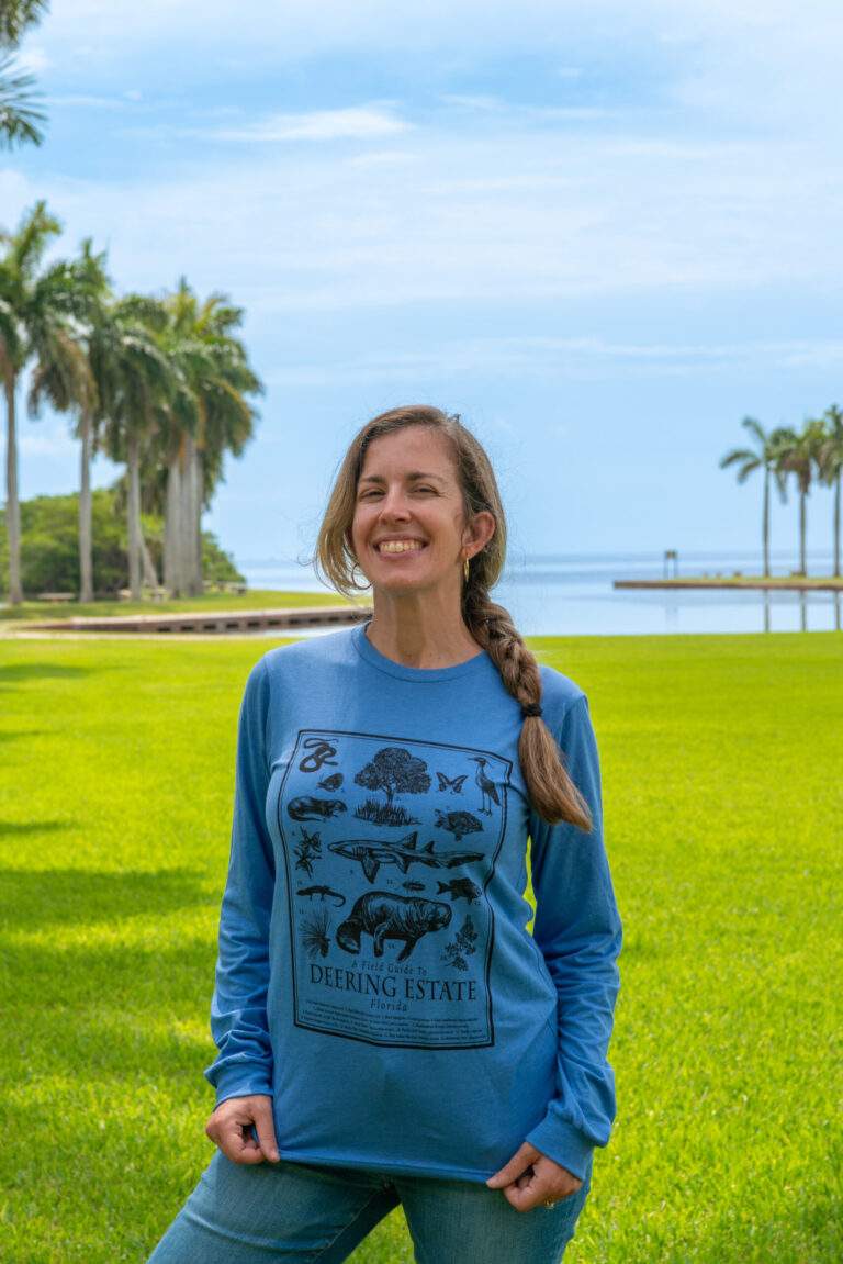 A person stands in front of the Main Lawn showing off their t-shirt, which features a design of a Deering Estate Field Guide.